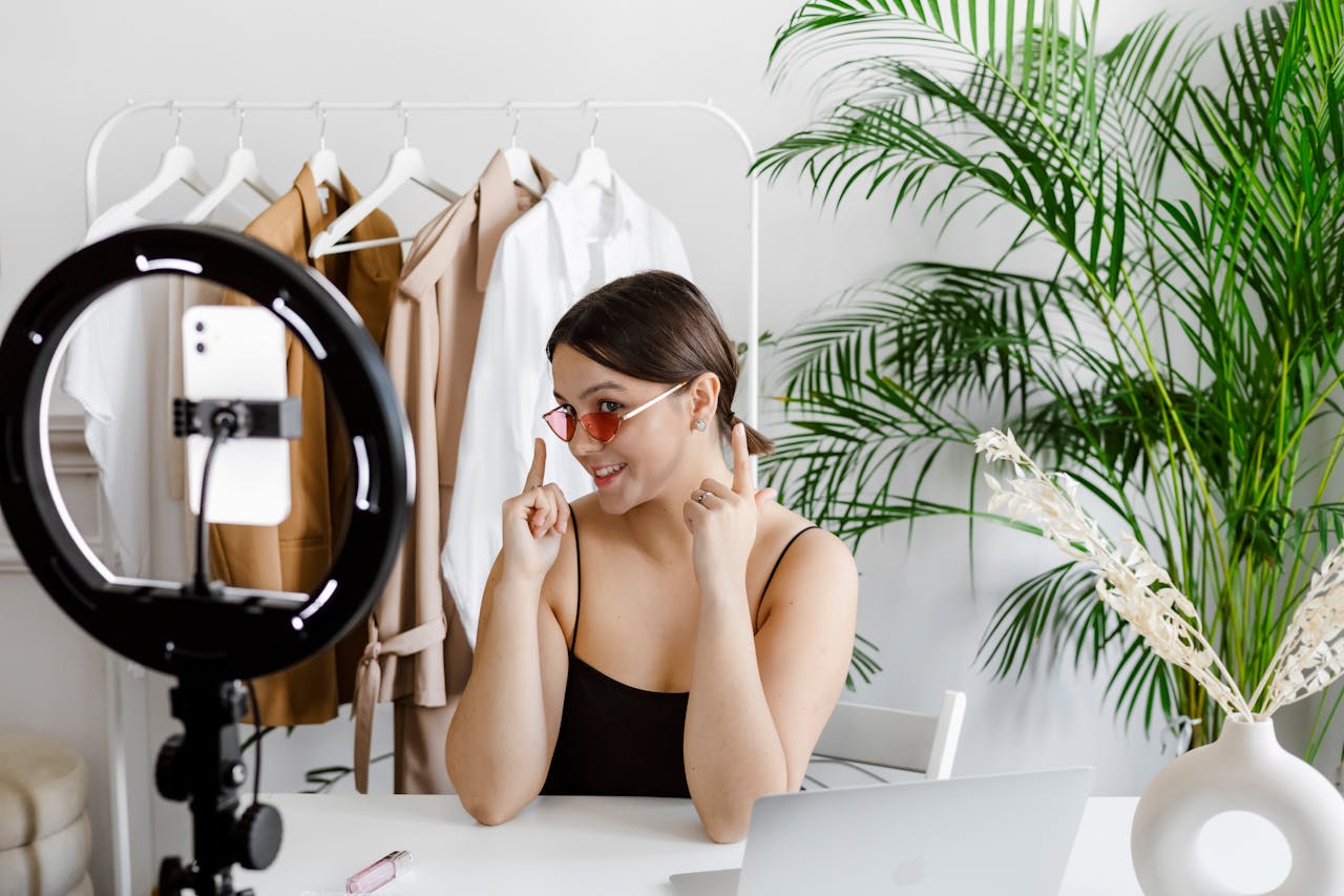 gallery-04 Stylish woman vlogging with ring light, surrounded by fashion accessories and greenery.
